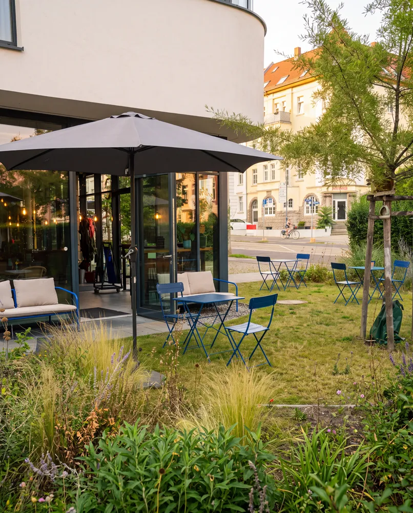 Sommerlicher Außenbereich des kate Cafés mit blauen Bistrotischen, Sonnenschirm und Garten